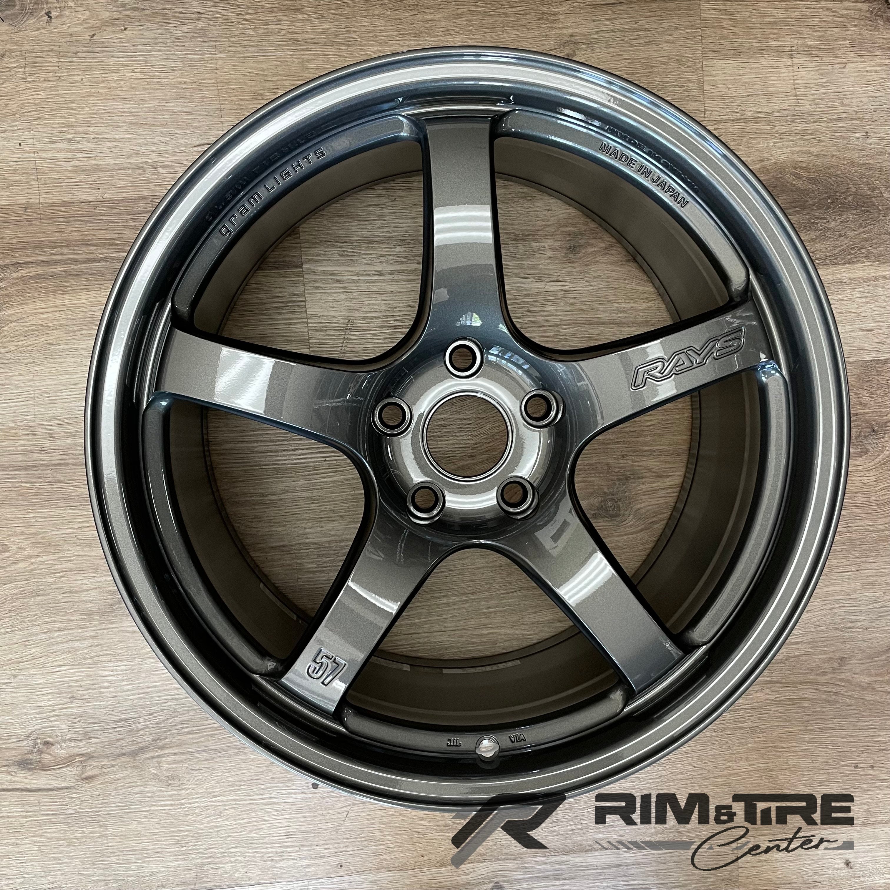 Photograph of a RAYS Gram Lights 57 five-spoke dark metallic gray car wheel with 'Made in Japan' marking, displayed on a light wooden floor with a 'Rim & Tire Center' logo in the bottom right corner