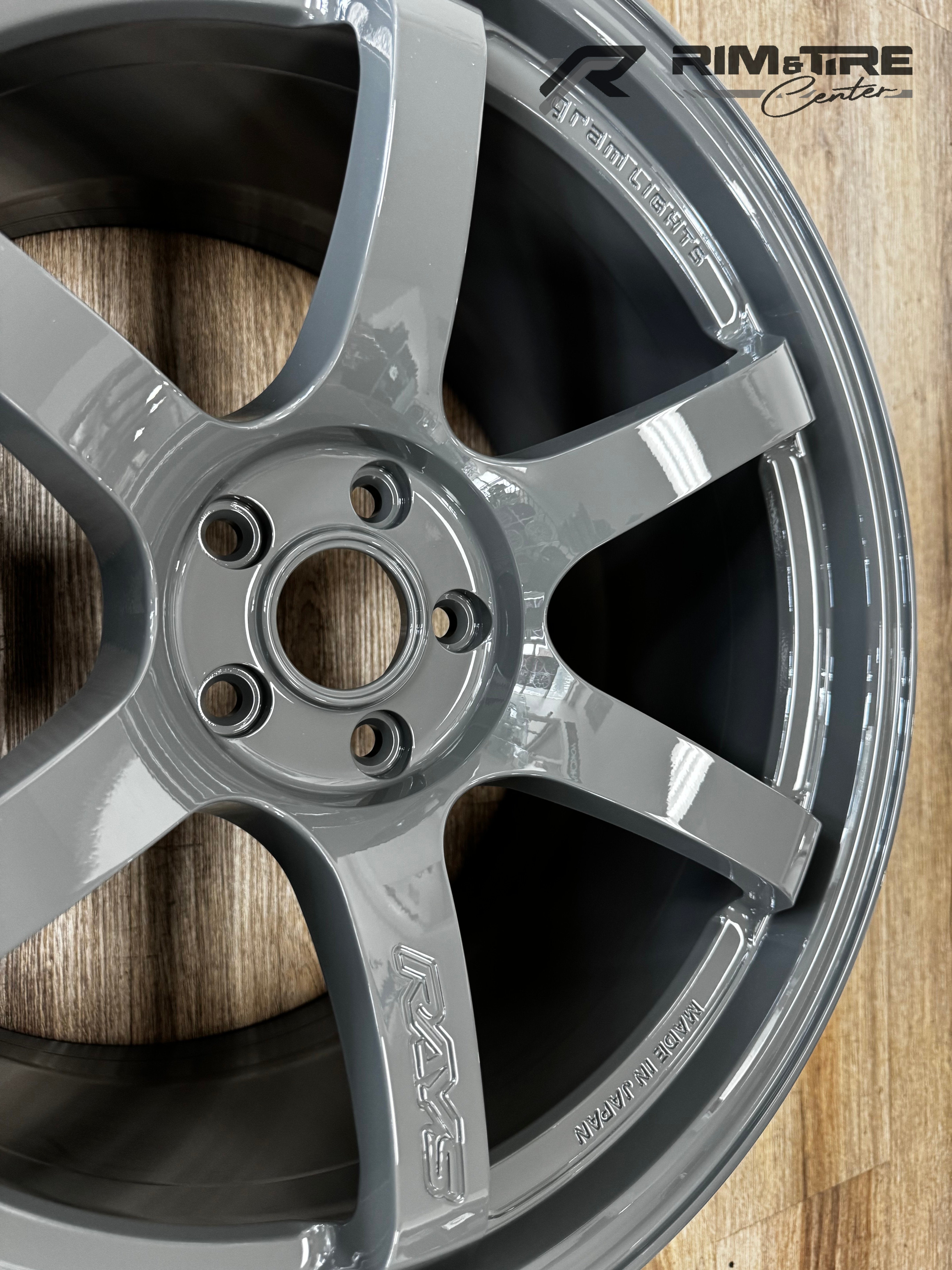 Product photograph of a glossy light grey RAYS Gram Lights five-spoke automotive rim with MADE IN JAPAN inscription resting on a light brown wooden floor at a RIM & TIRE Center