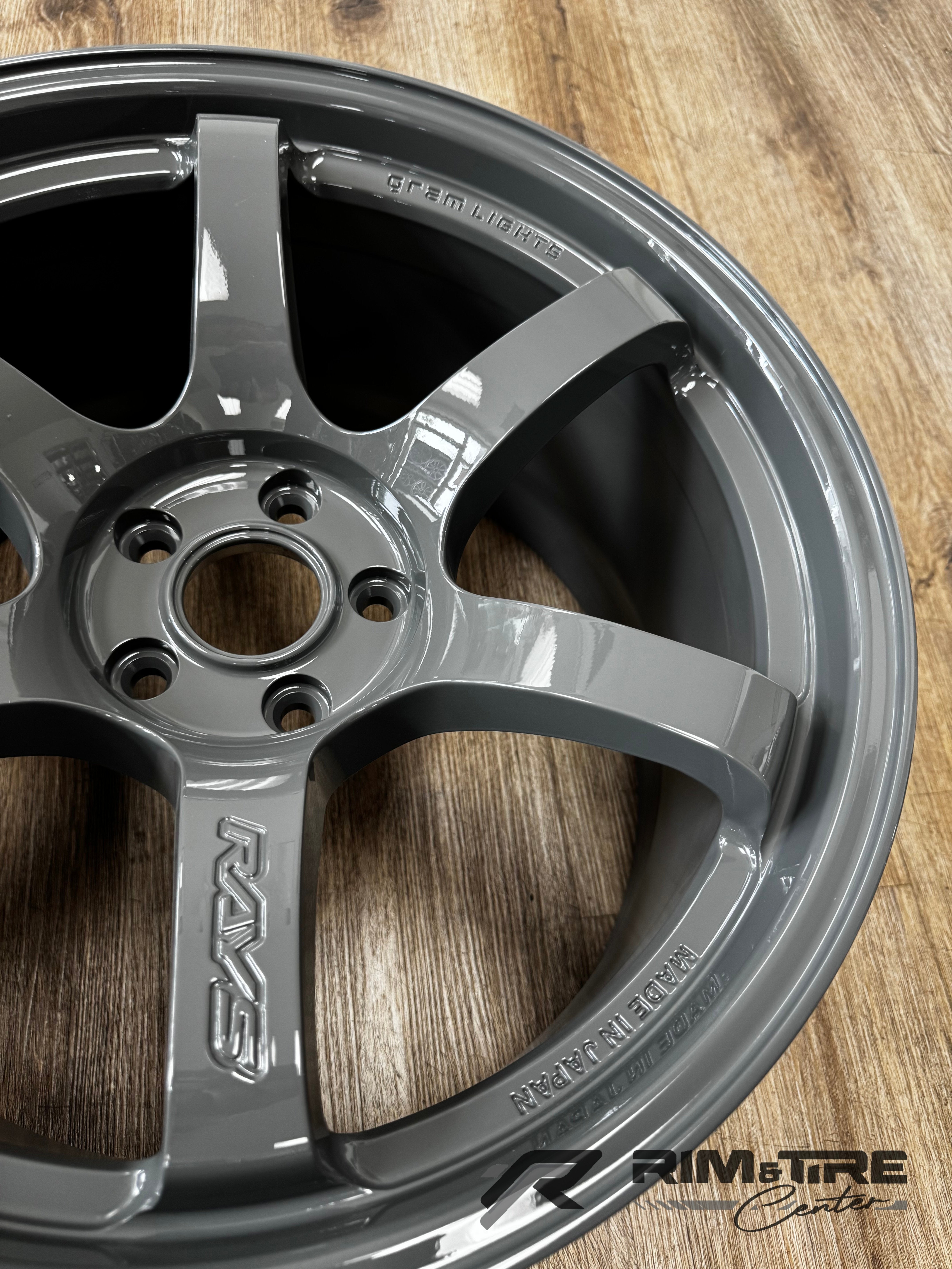Photograph of a RAYS Gram Lights five-spoke automotive wheel in glossy gunmetal gray finish featuring embossed RAYS and MADE IN JAPAN lettering displayed on a light brown wooden floor at Rim & Tire Center