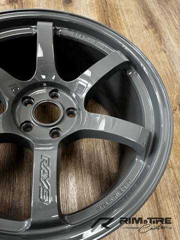 Photograph of a RAYS Gram Lights five-spoke automotive wheel in glossy gunmetal gray finish featuring embossed RAYS and MADE IN JAPAN lettering displayed on a light brown wooden floor at Rim & Tire Center