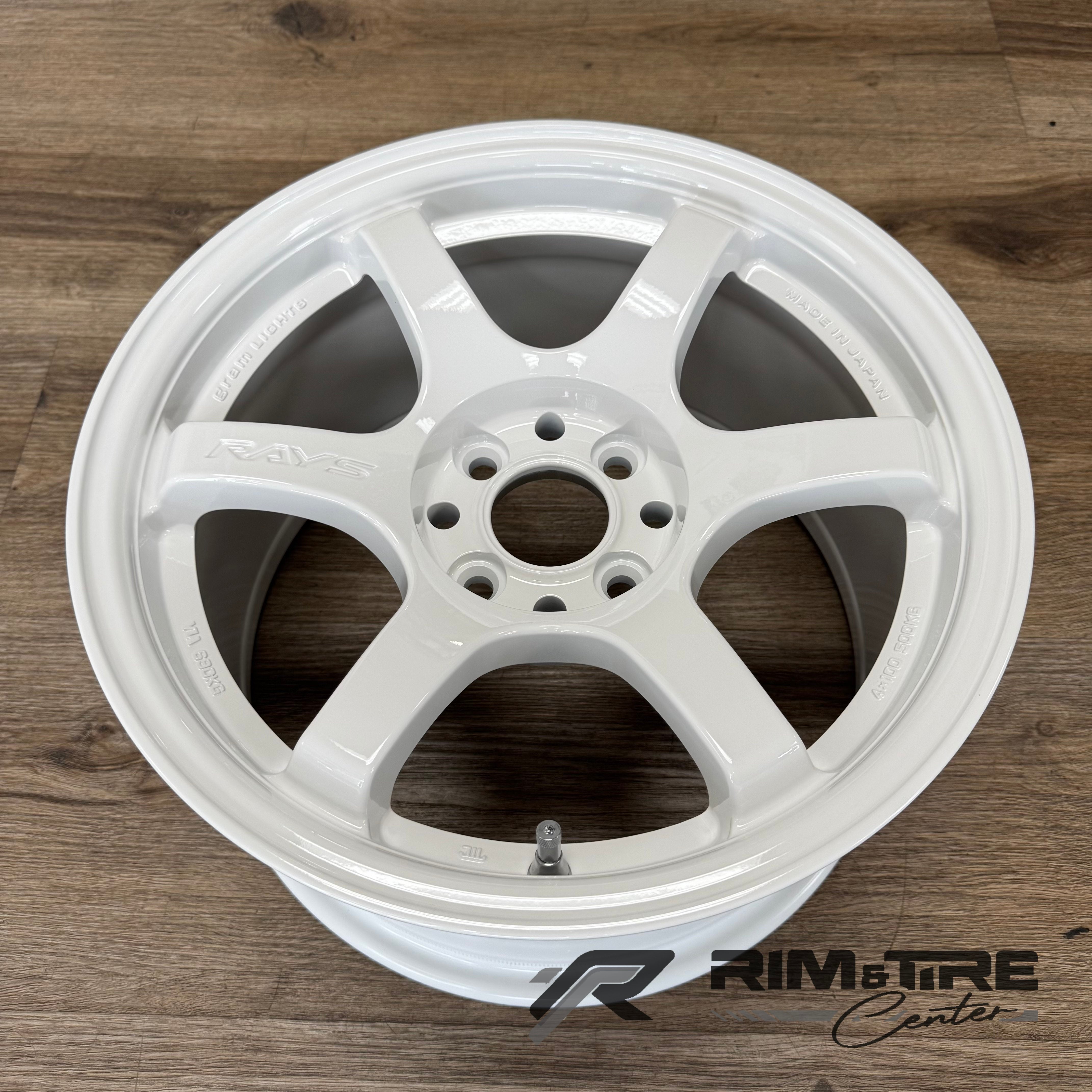 Product photography of a glossy white RAYS Gram Lights 57CR 6-spoke car wheel with embossed 'RAYS', 'GRAM LIGHTS', and 'MADE IN JAPAN' text, featuring a chrome valve stem, resting on a brown wooden floor