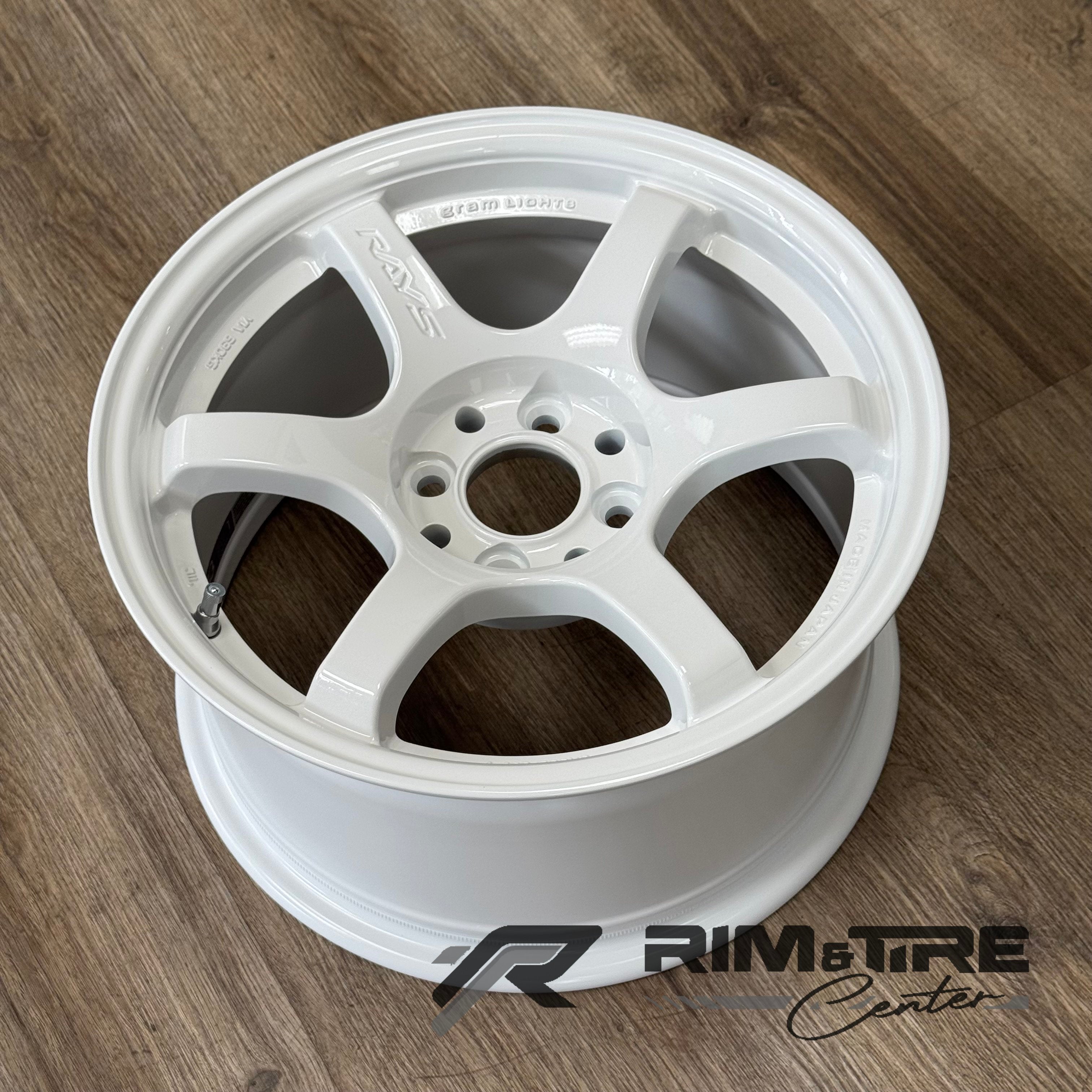 Photograph of a RAYS Gram Lights 57DR six-spoke alloy wheel in glossy white finish with a visible valvestem on a warm brown wood grain floor