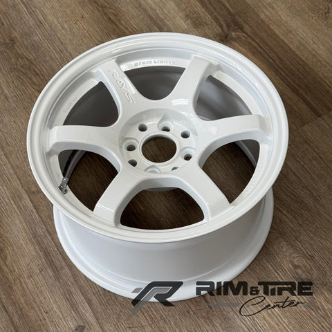 Photograph of a RAYS Gram Lights 57DR six-spoke alloy wheel in glossy white finish with a visible valvestem on a warm brown wood grain floor