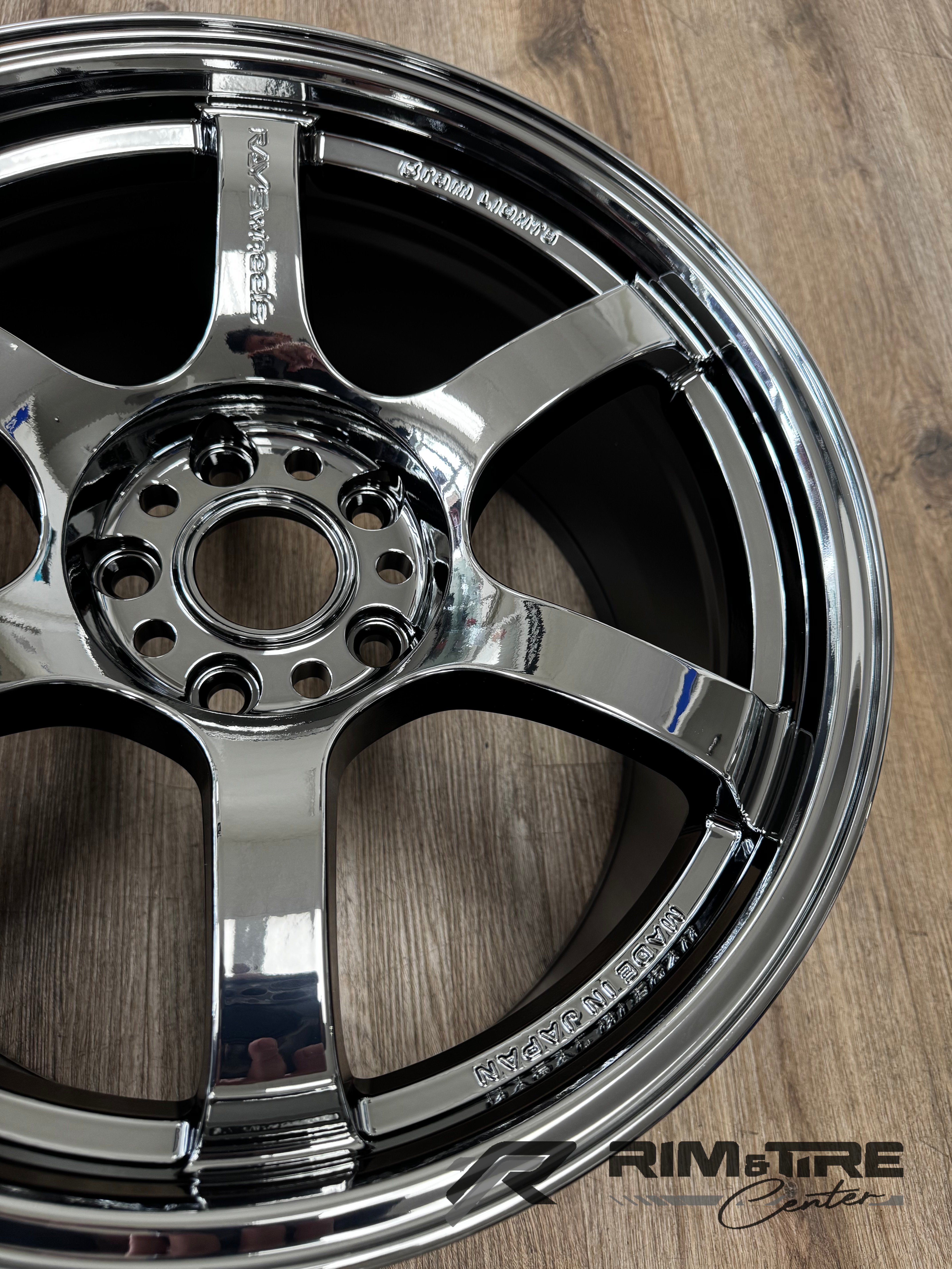 Photograph of a highly reflective chrome RAYS Wheels five-spoke car wheel featuring a multi-lug bolt pattern and "MADE IN JAPAN" engraving, resting on a textured light brown wooden floor