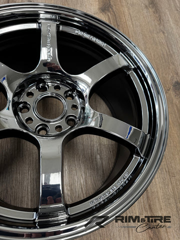 Photograph of a highly reflective chrome RAYS Wheels five-spoke car wheel featuring a multi-lug bolt pattern and "MADE IN JAPAN" engraving, resting on a textured light brown wooden floor