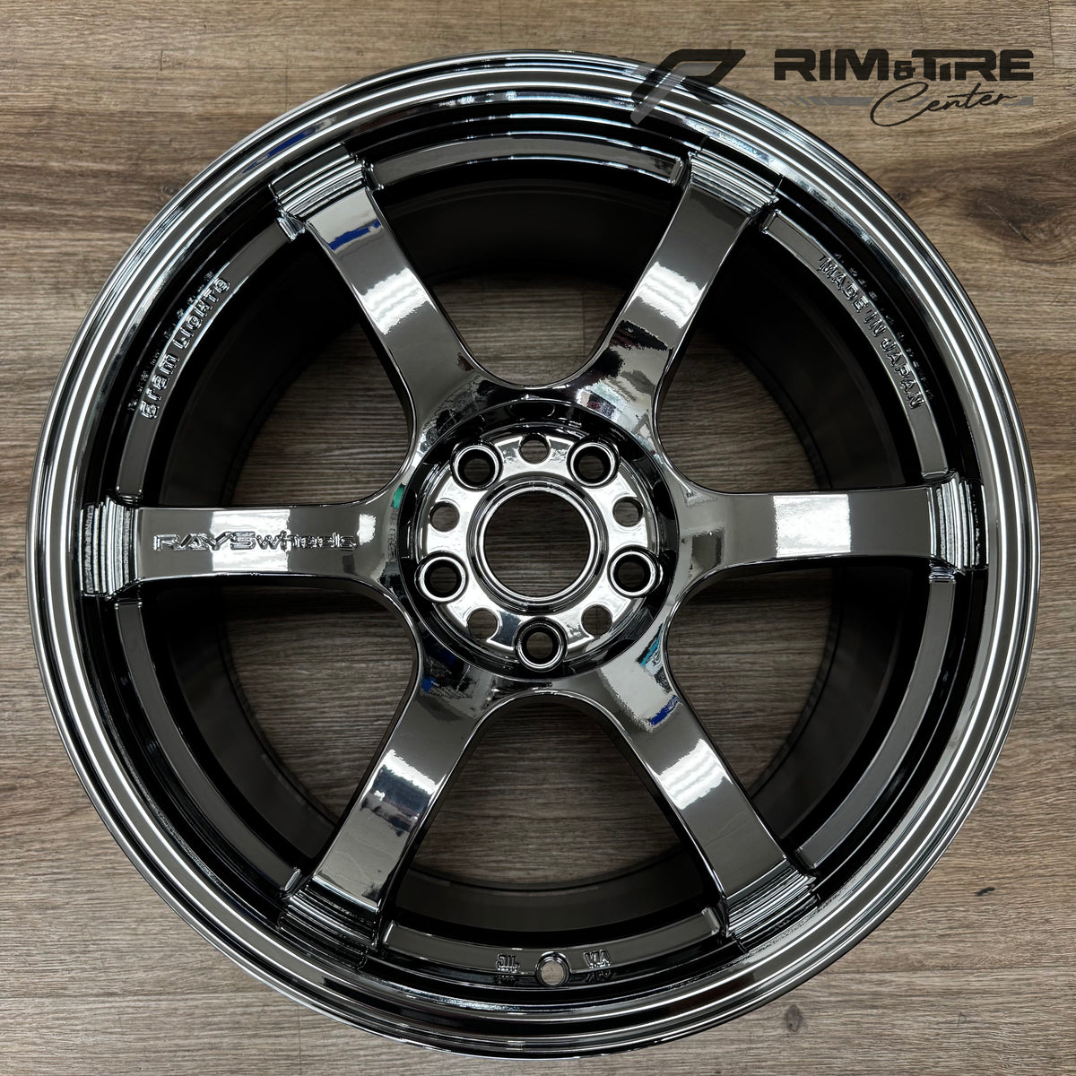 Digital photograph of a RAYSWheels Gram Lights 57CR 6-spoke alloy wheel in glossy black chrome finish, displaying 'Made in Japan' engraving, positioned on a light brown wooden floor with a 'RIM & TIRE Center' watermark