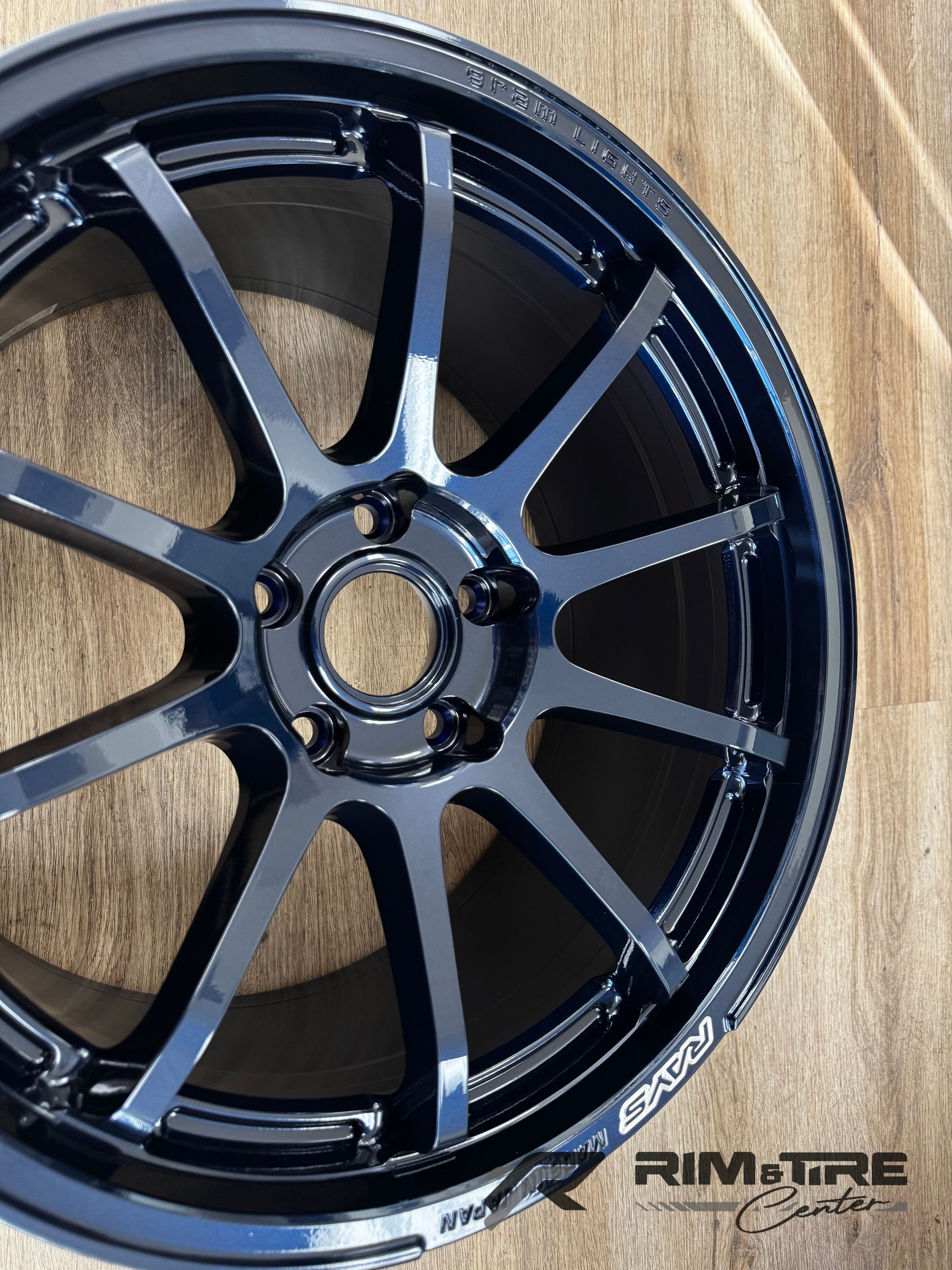 Photograph of a dark metallic blue RAYS Gram Lights multi-spoke alloy wheel with glossy finish, showing Made in Japan text on a wooden floor with a Rim & Tire Center watermark