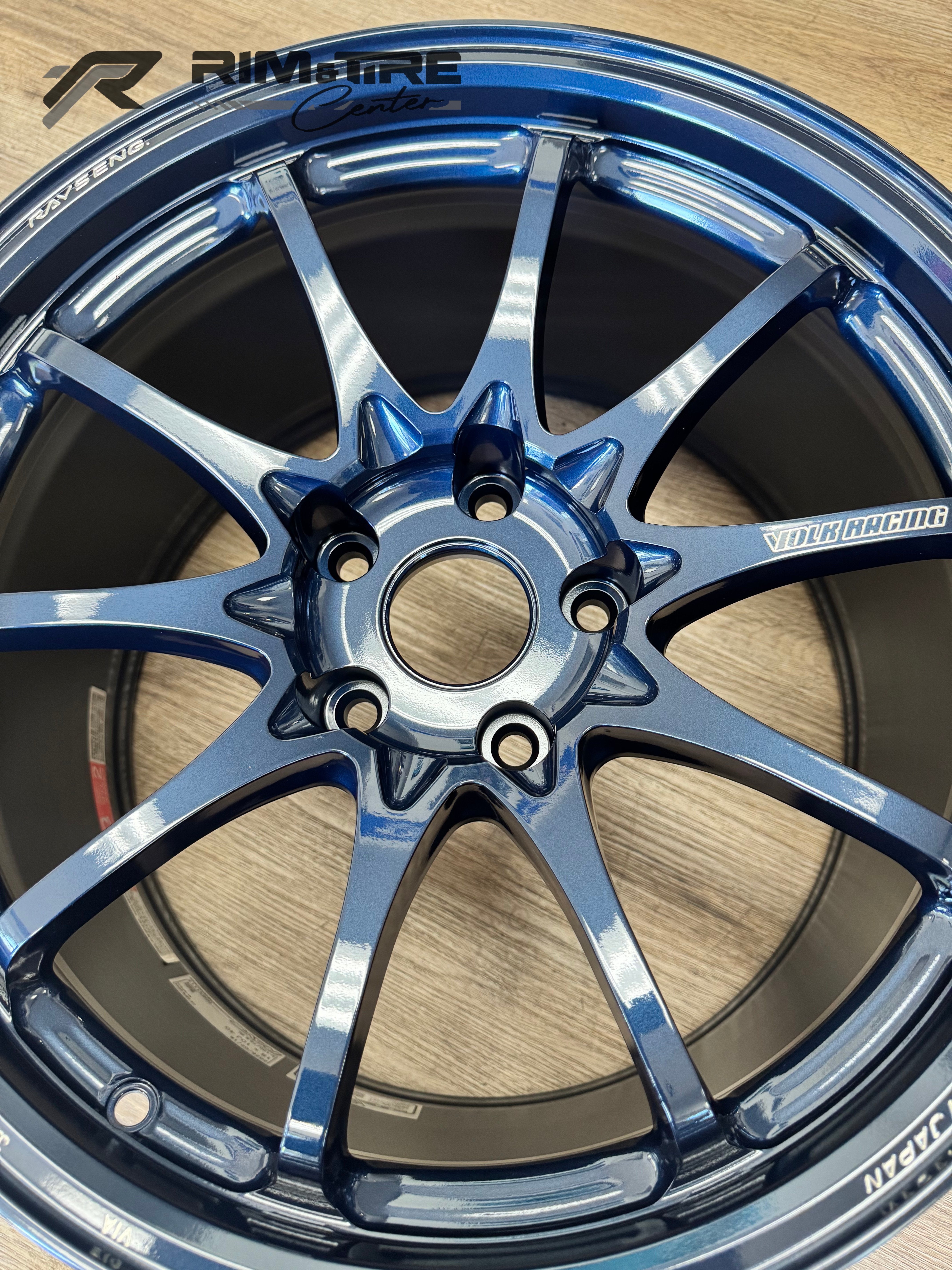 Automotive photography of a RAYSENG / VOLK RACING dark blue metallic multi-spoke car wheel on a wooden surface, showcasing its glossy finish, intricate 6-lug pattern, and "MADE IN JAPAN" branding