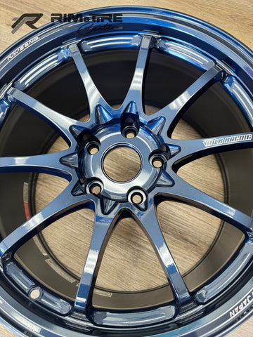 Automotive photography of a RAYSENG / VOLK RACING dark blue metallic multi-spoke car wheel on a wooden surface, showcasing its glossy finish, intricate 6-lug pattern, and "MADE IN JAPAN" branding