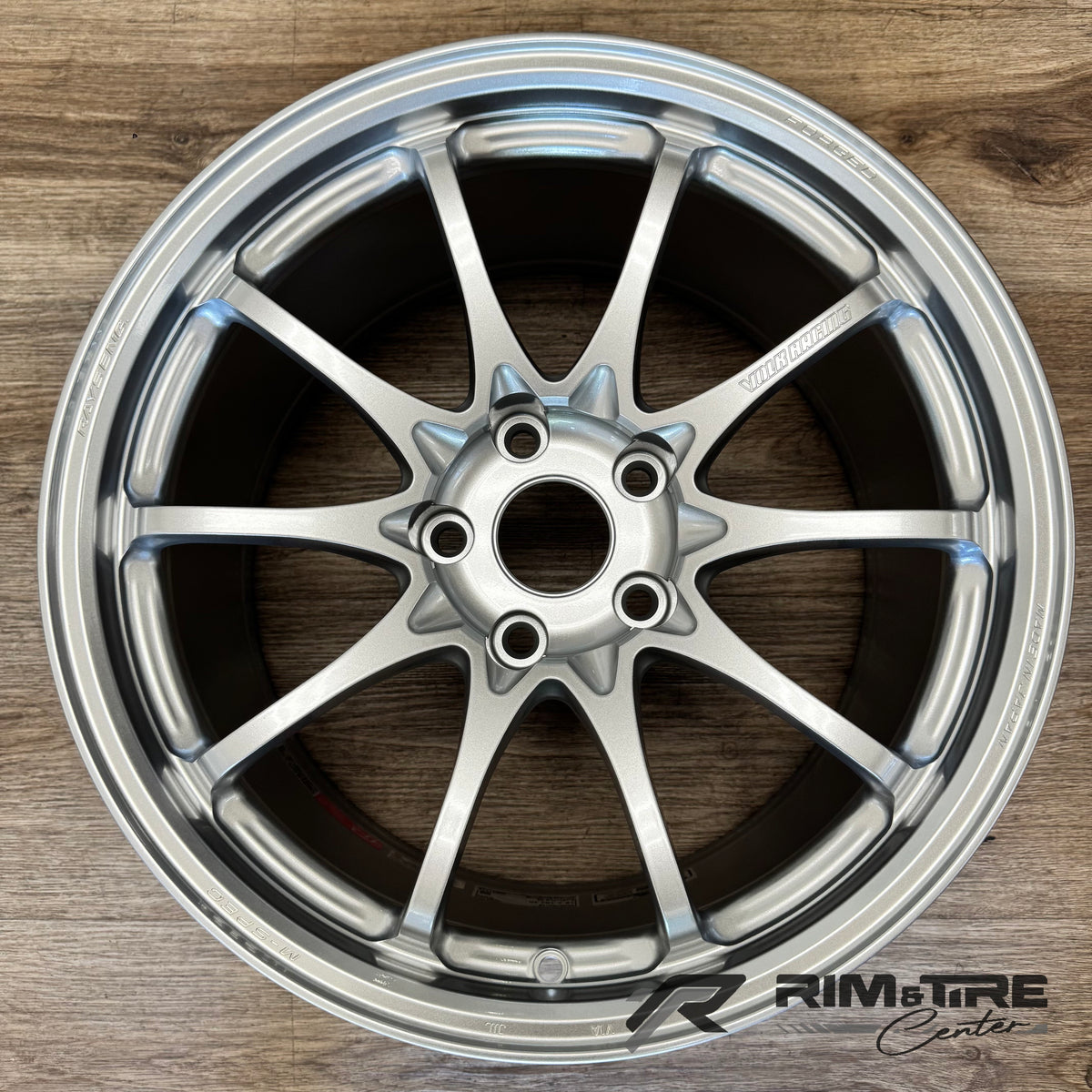Product photo by Rim & Tire Center of a shiny silver 10-spoke Volk Racing forged car wheel Made in Japan displayed on a textured wooden floor