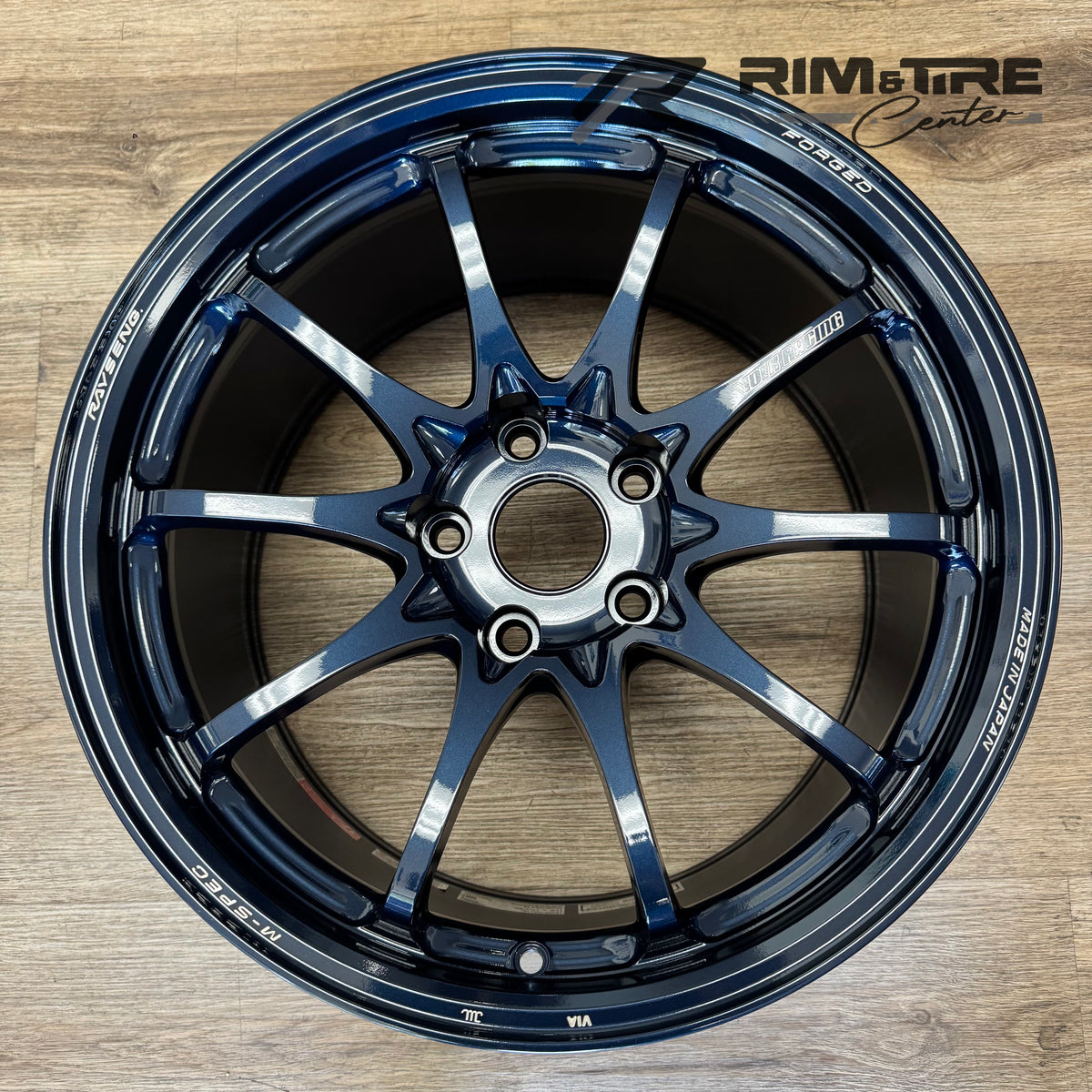 Photograph of a RAYS VOLK RACING FORGED M-SPEC ten-spoke car wheel in a vibrant metallic dark blue finish with "MADE IN JAPAN" text visible, presented on a textured wooden floor