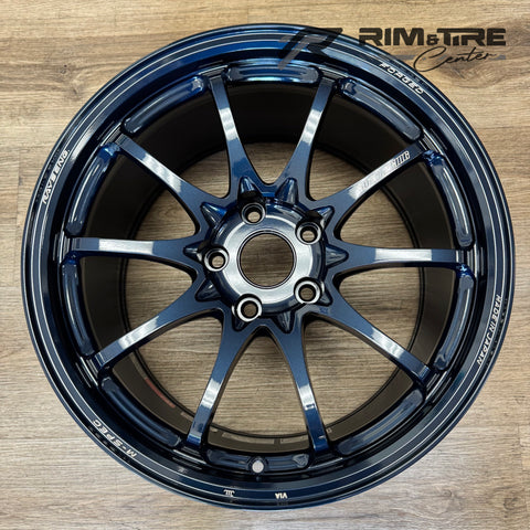 Photograph of a RAYS VOLK RACING FORGED M-SPEC ten-spoke car wheel in a vibrant metallic dark blue finish with "MADE IN JAPAN" text visible, presented on a textured wooden floor