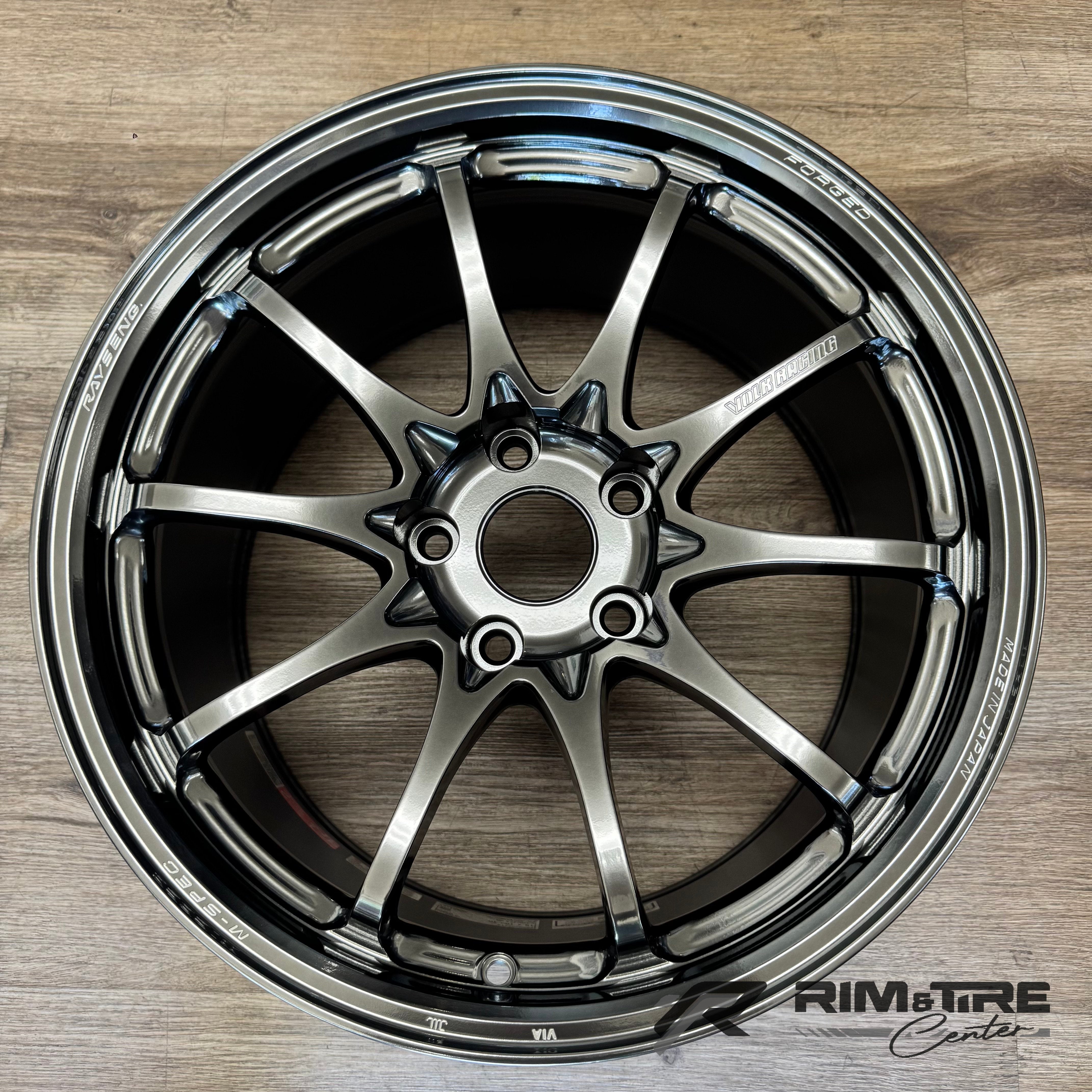 Photograph of a single dark chrome ten-spoke car wheel by Volk Racing Rays Engineering, marked FORGED and MADE IN JAPAN, sitting on a light wooden floor displaying its metallic gunmetal finish and detailed center hub