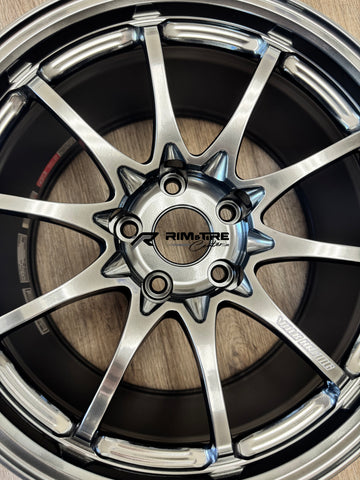 Photograph of a Volk Racing multi-spoke automotive rim with metallic silver finish and glossy black details displaying a Rim&Tire Center logo spiked lug covers and a red accent sticker on a wooden floor