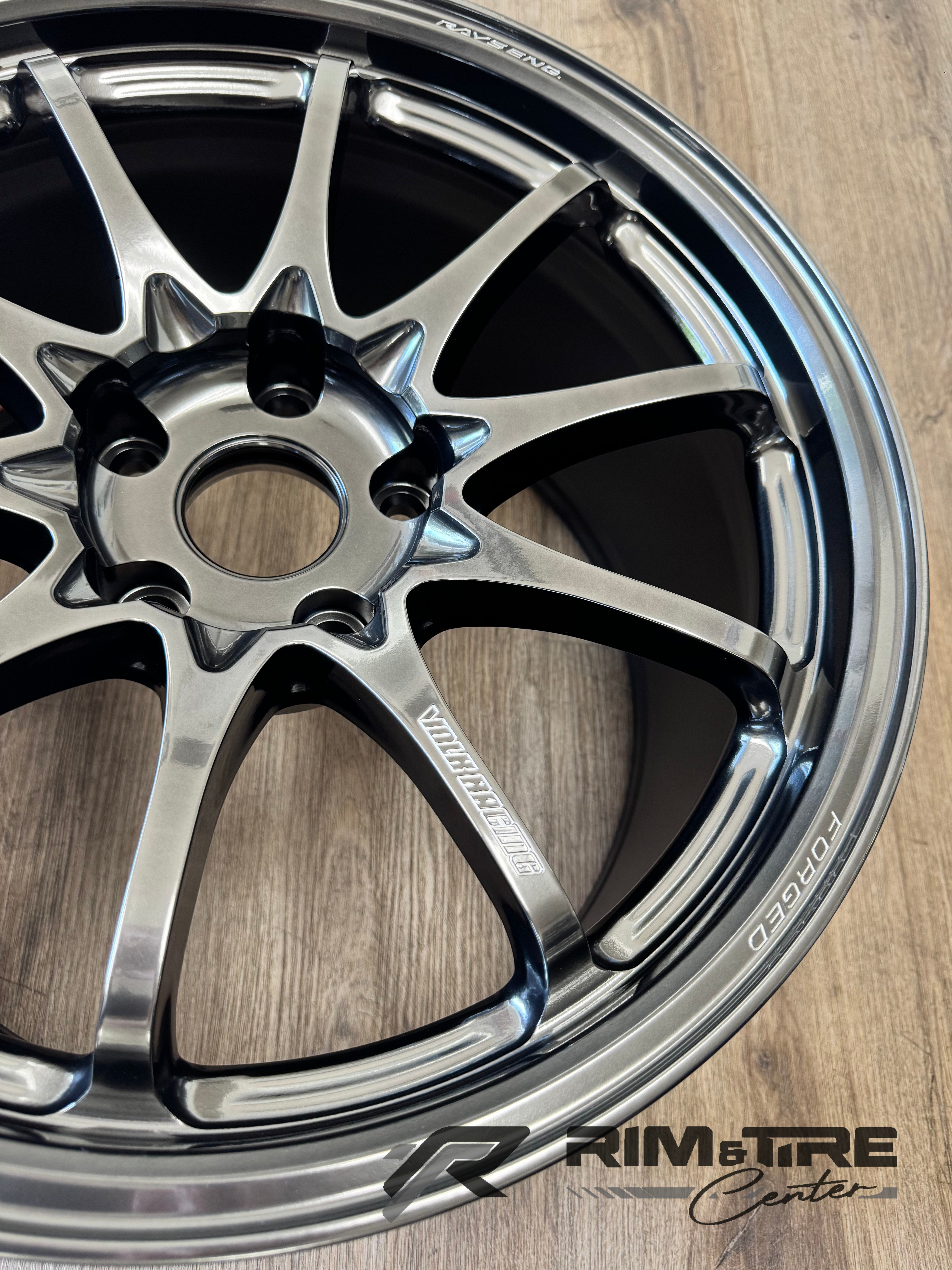Product photography of a RAYS ENG Volk Racing forged multi-spoke wheel featuring a glossy dark chrome finish and prominent FORGED inscription, displayed on a light brown wooden floor