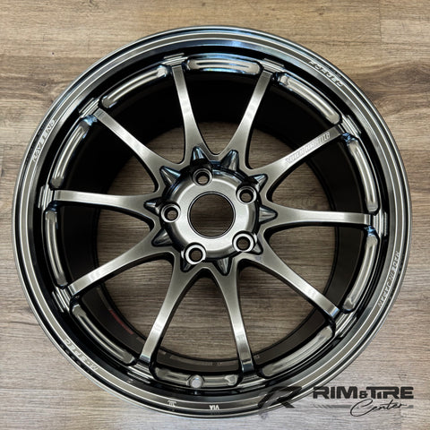 Photograph of a single dark chrome ten-spoke car wheel by Volk Racing Rays Engineering, marked FORGED and MADE IN JAPAN, sitting on a light wooden floor displaying its metallic gunmetal finish and detailed center hub