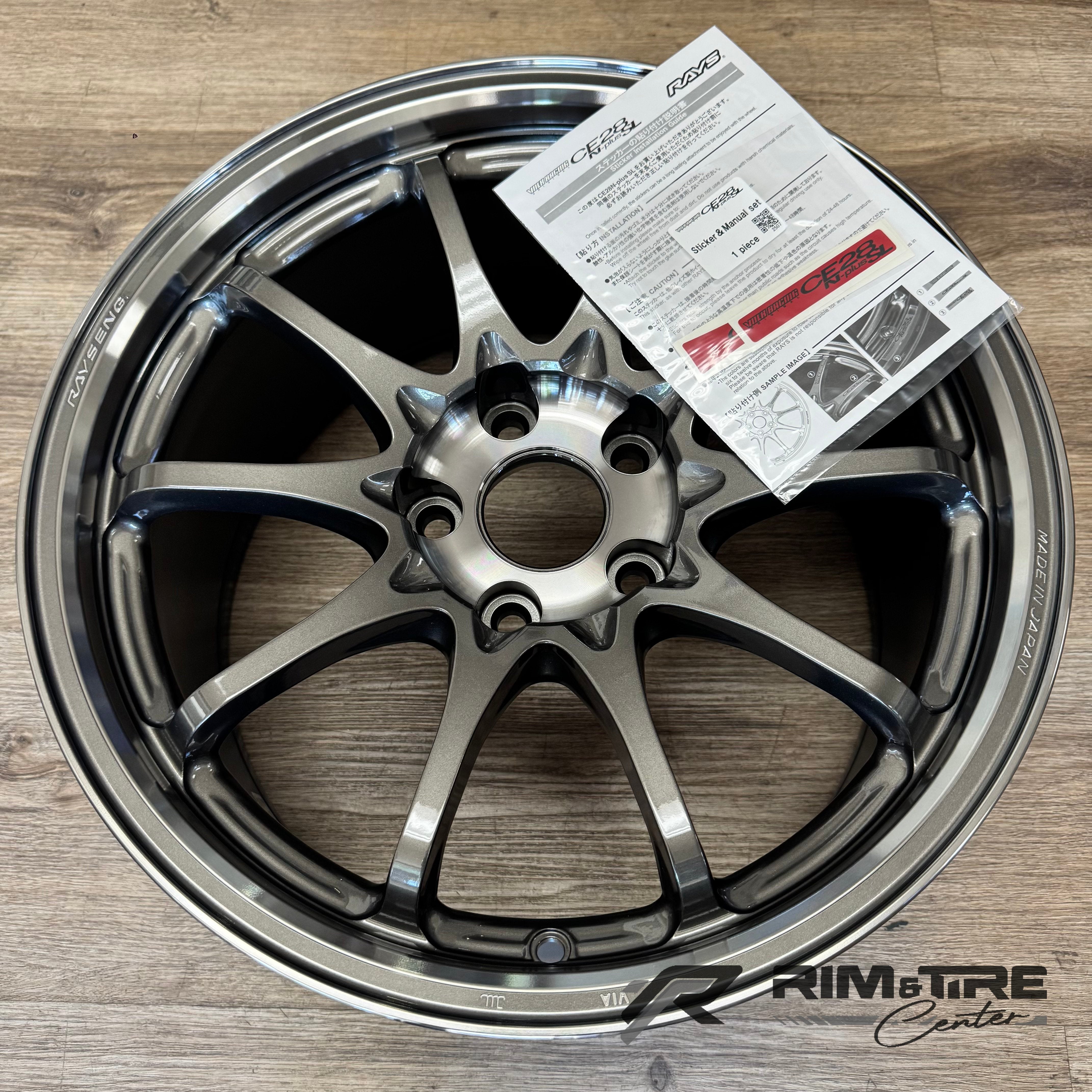 Photograph of a RAYS Engineering Volk Racing CE28N-plus SL 10-spoke alloy wheel in dark metallic grey on a light wood floor, featuring "RAYS ENG." and "MADE IN JAPAN" etched details and an accompanying RAYS sticker installation guide