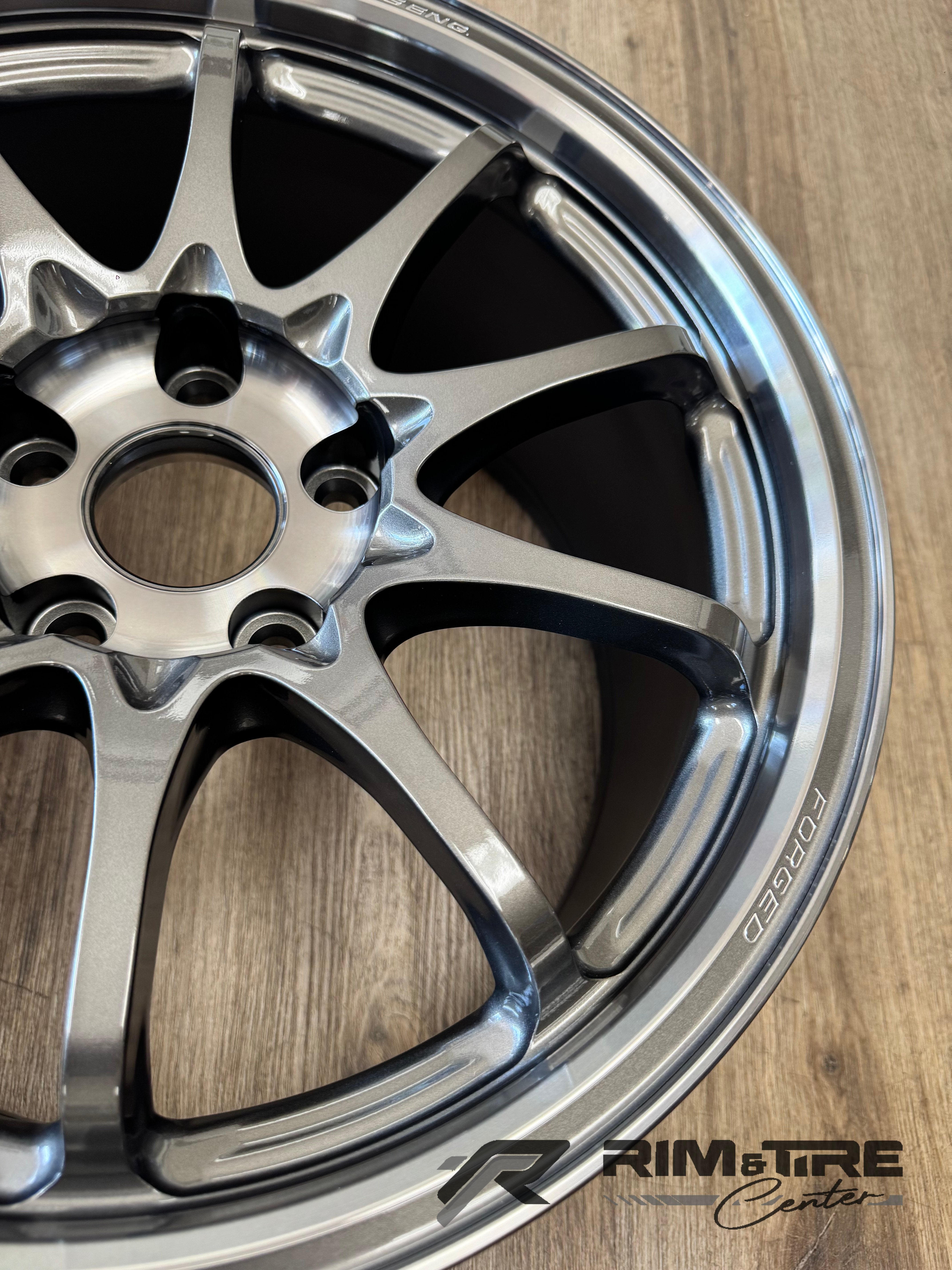 Photograph by RIM&TIRE Center of a glossy gunmetal multi-spoke forged car wheel with 'FORGED' text on its rim, positioned on a light brown wooden floor