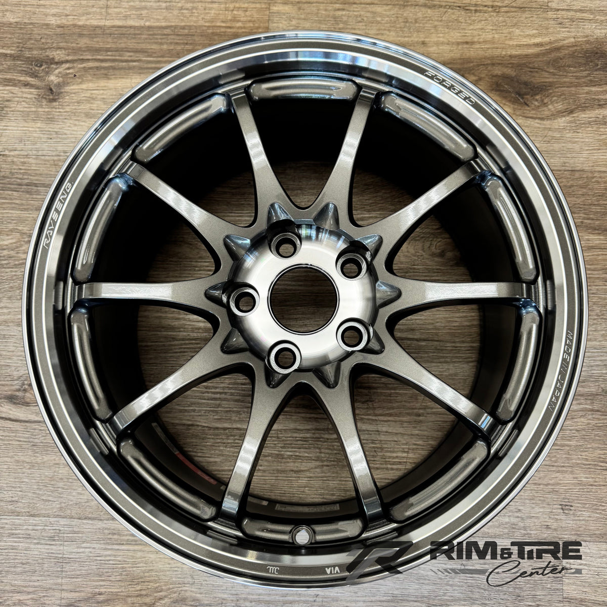 Photograph of a Rays Engineering forged gunmetal 10-spoke car wheel featuring a polished lip and 'RAYS ENG MADE IN JAPAN' branding, resting on a light wood floor