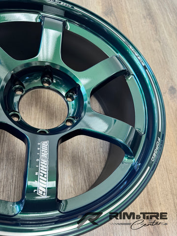 Product photo of a shiny iridescent dark metallic green-blue Volk Racing Original 6-spoke forged car wheel rim with prominent 'VOLK RACING' and 'FORGED' branding on a warm wooden floor