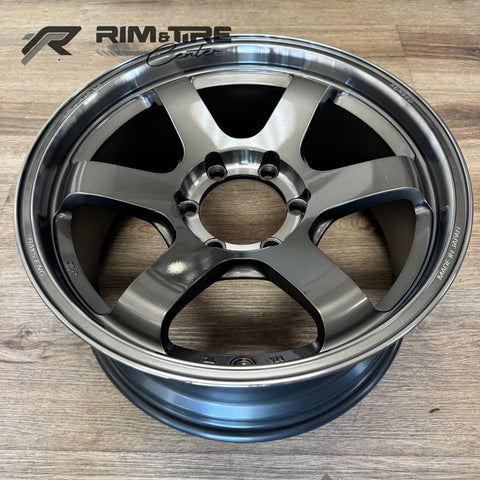 Photograph of a RAYS ENG. Volk Racing forged car wheel in dark metallic gray featuring a prominent five-spoke design, a bright polished lip, and visible "MADE IN JAPAN" text on a warm wooden floor
