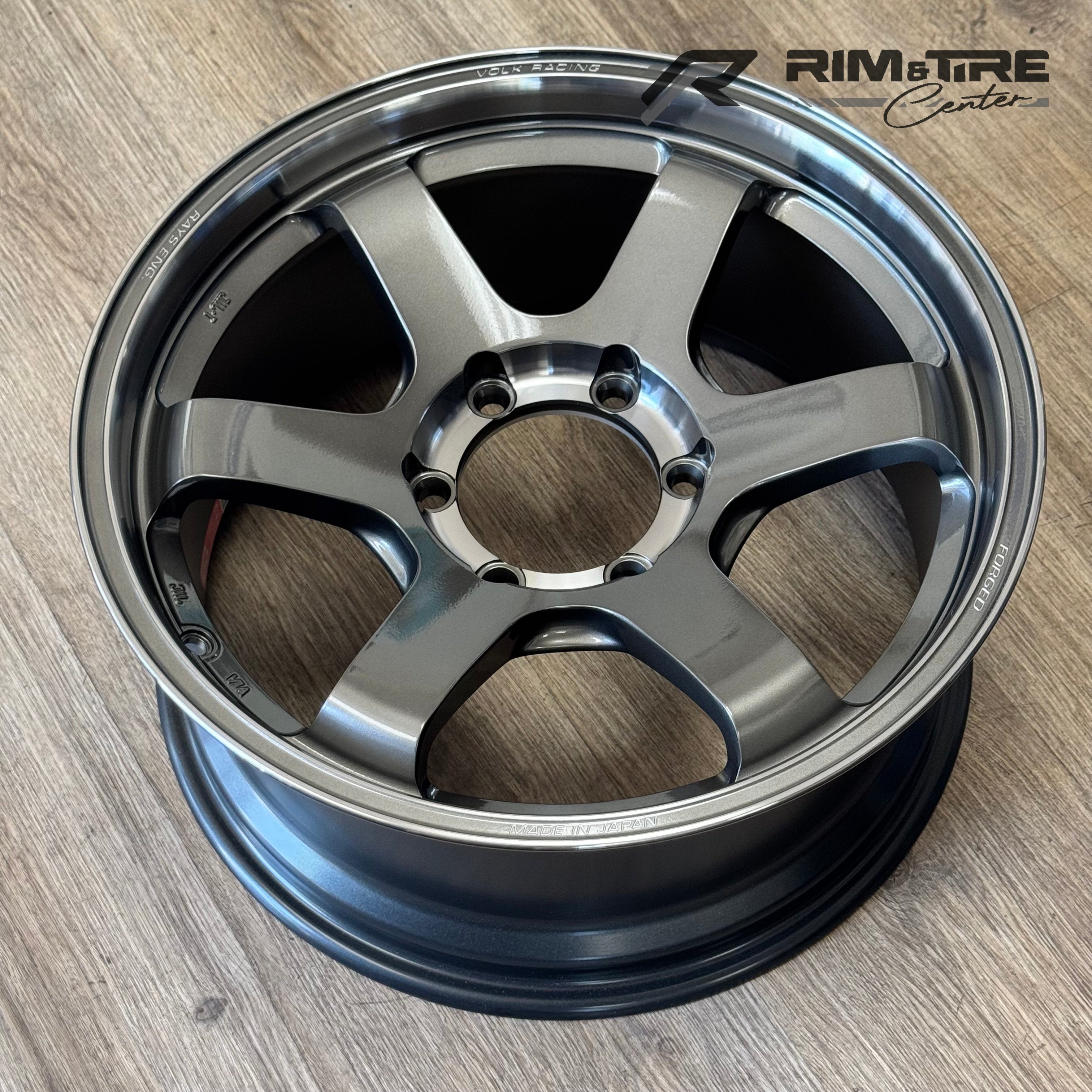 Photography of a RAYS Engineering Volk Racing TE37 SL forged wheel in a metallic Pressed Graphite finish with a distinct six-spoke design and polished outer lip, highlighting its 'MADE IN JAPAN' quality, on a light wooden floor