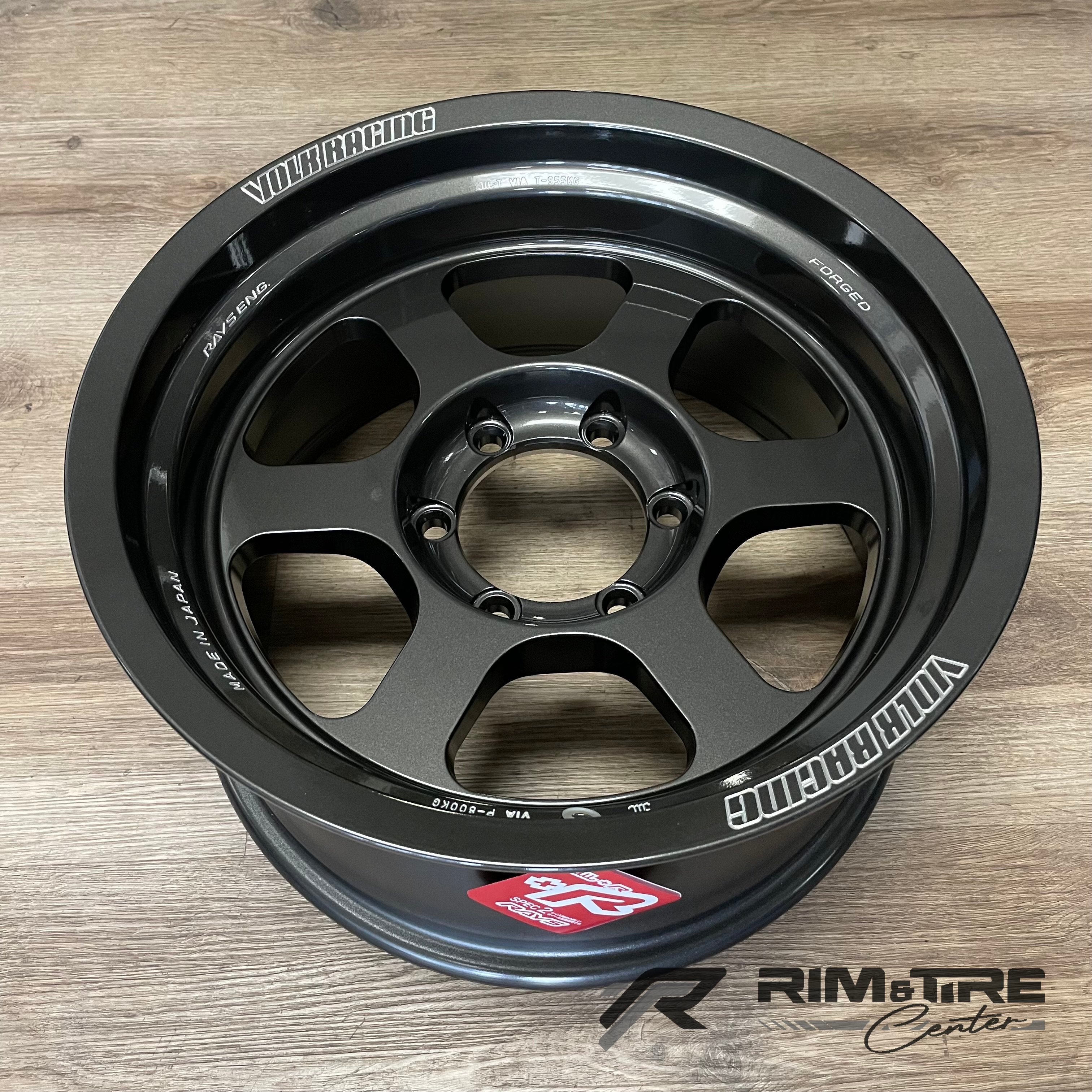 Product photograph of a dark grey five-spoke Volk Racing Rays Engineering forged wheel with white branding and a red RAYS sticker resting on a light wood floor