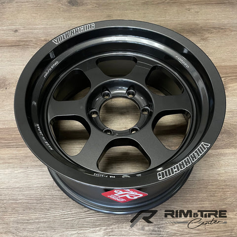 Product photograph of a dark grey five-spoke Volk Racing Rays Engineering forged wheel with white branding and a red RAYS sticker resting on a light wood floor