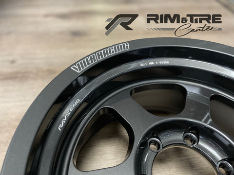 Photography of a matte dark grey VOLK RACING wheel rim with RAYS ENG and JWL-T VIA T-955KG markings on a light wooden surface, featuring the RIM & TIRE Center logo
