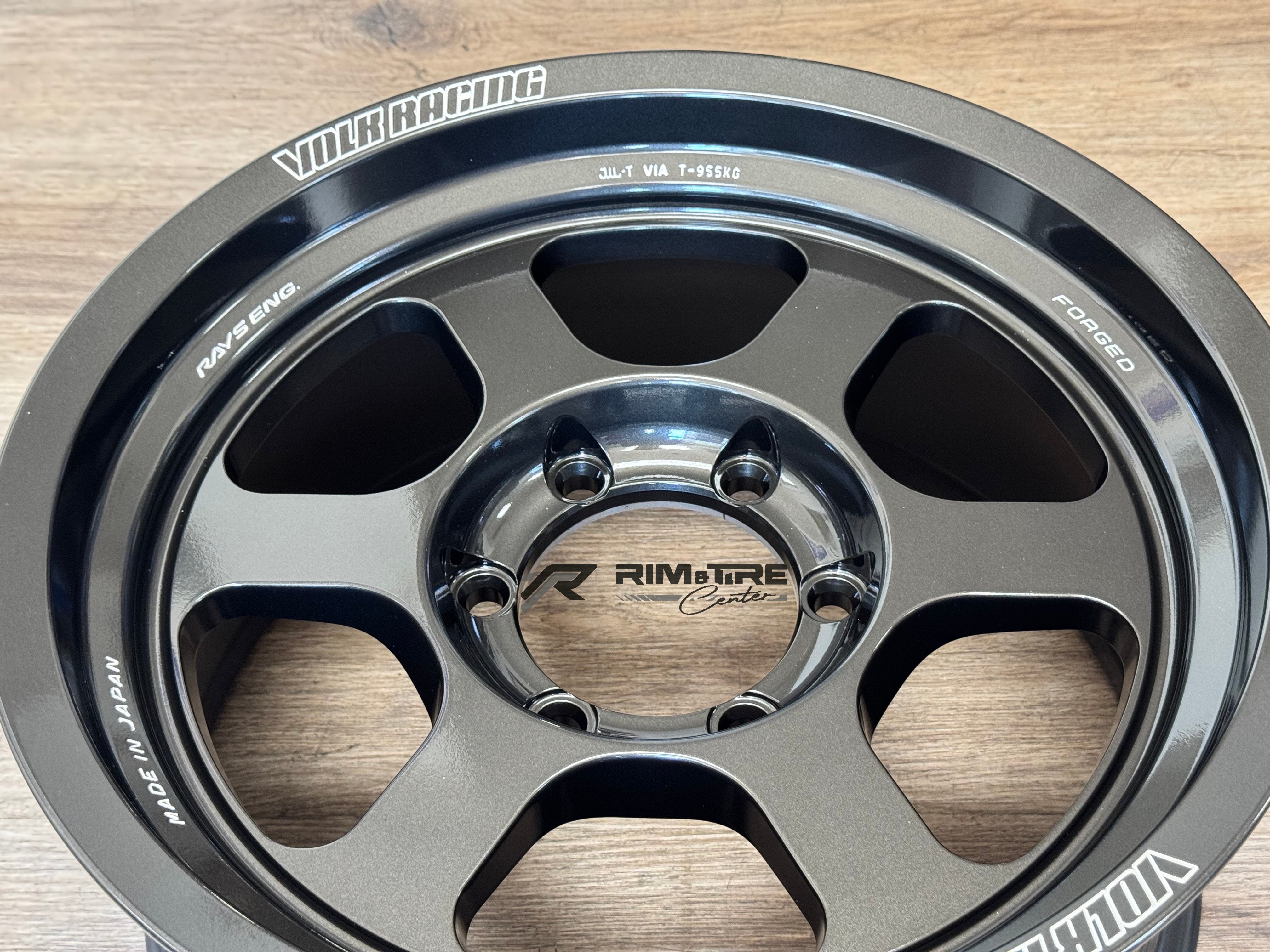 Photograph of a RAYS Engineering VOLK RACING dark gray forged five-spoke wheel featuring contrasting matte and glossy finishes, white branding, FORGED and MADE IN JAPAN text, presented on a light wooden surface