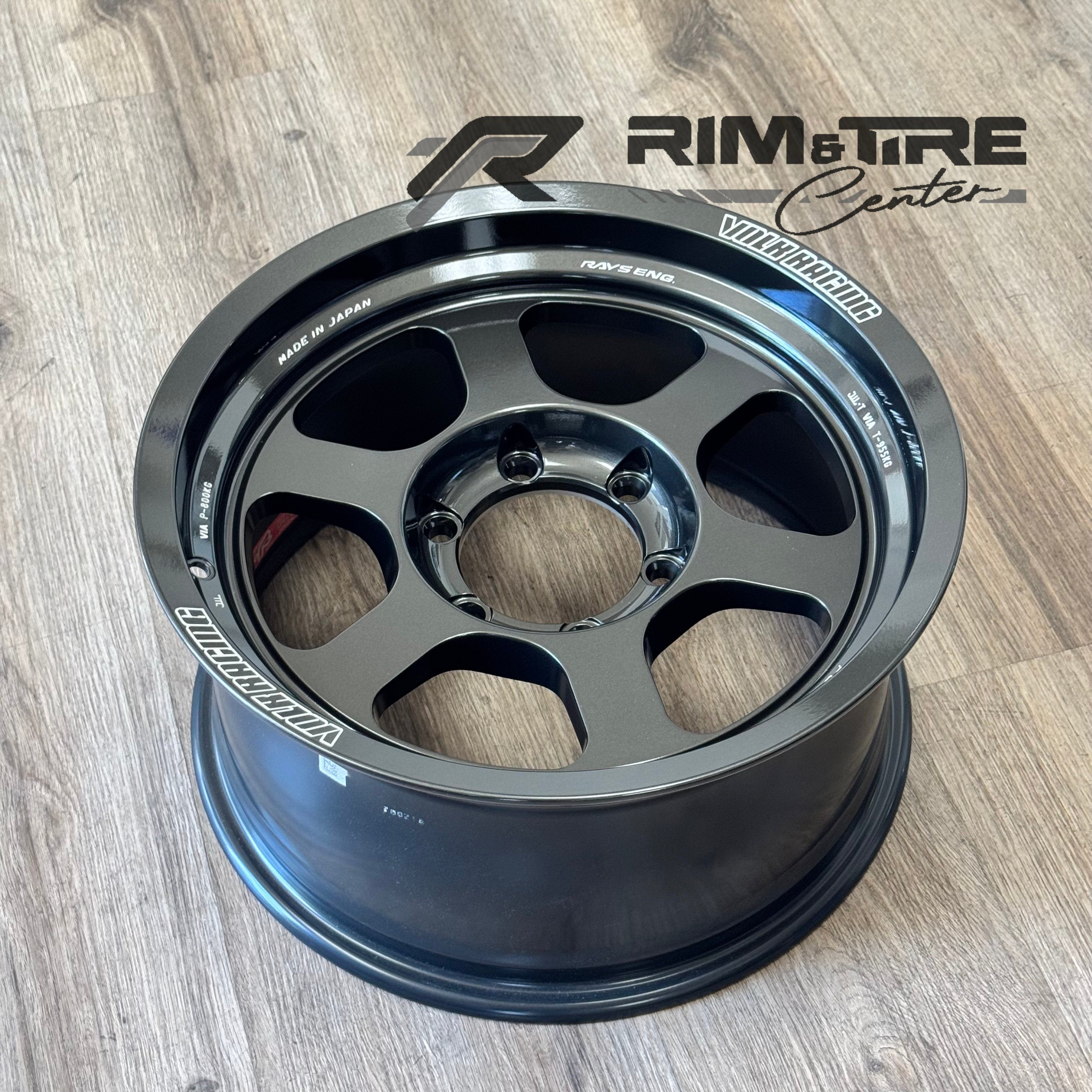 Digital photo of a dark metallic grey VOLK RACING RAYS ENG car wheel with six stylized cutouts and white text branding Made in Japan, presented on a light wood grain floor with a RIM & TIRE Center logo watermark