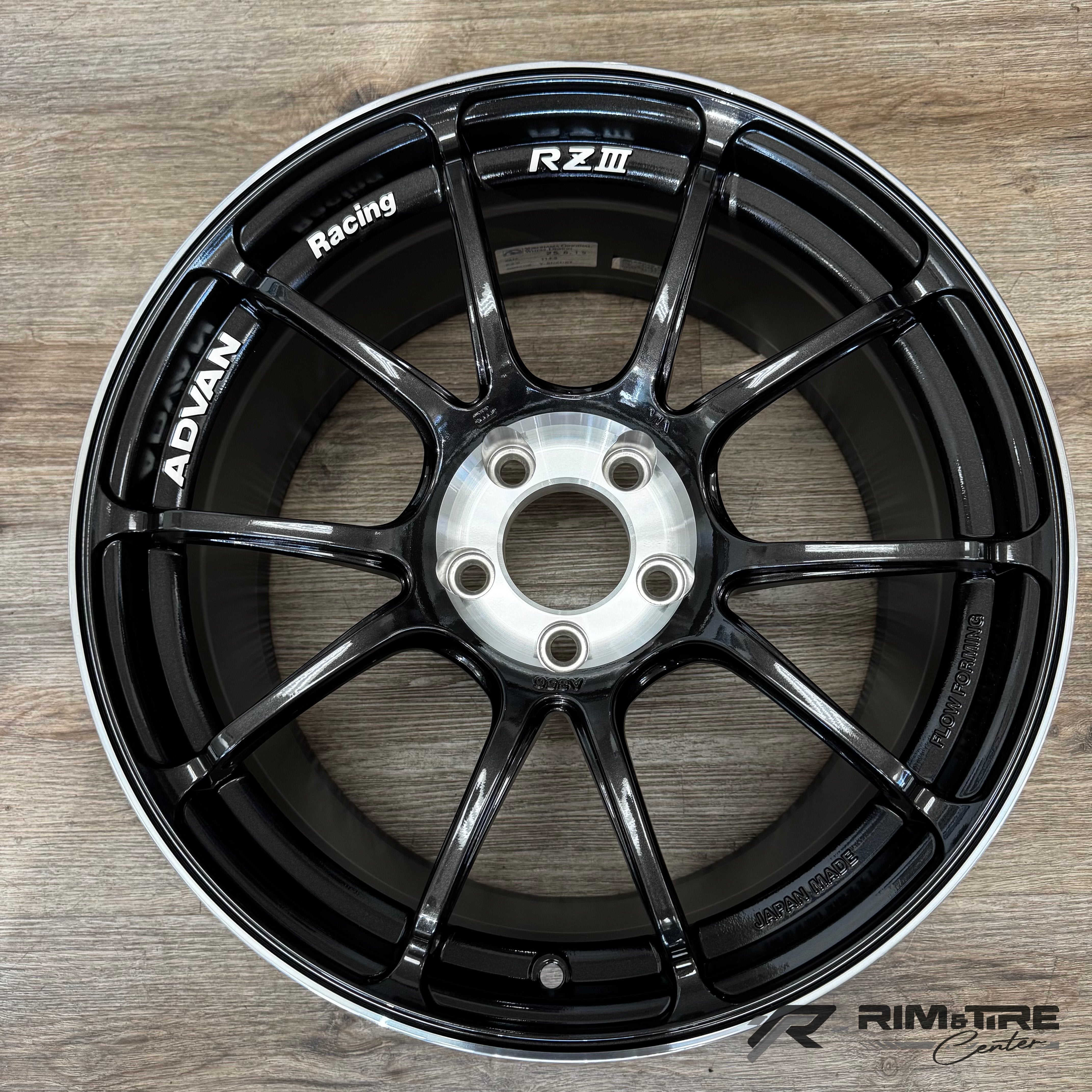 A glossy black Advan RZIII Racing multi-spoke car wheel with a silver lip and central hub, displaying 'ADVAN Racing RZIII' and 'JAPAN MADE' text, on a light brown wooden floor