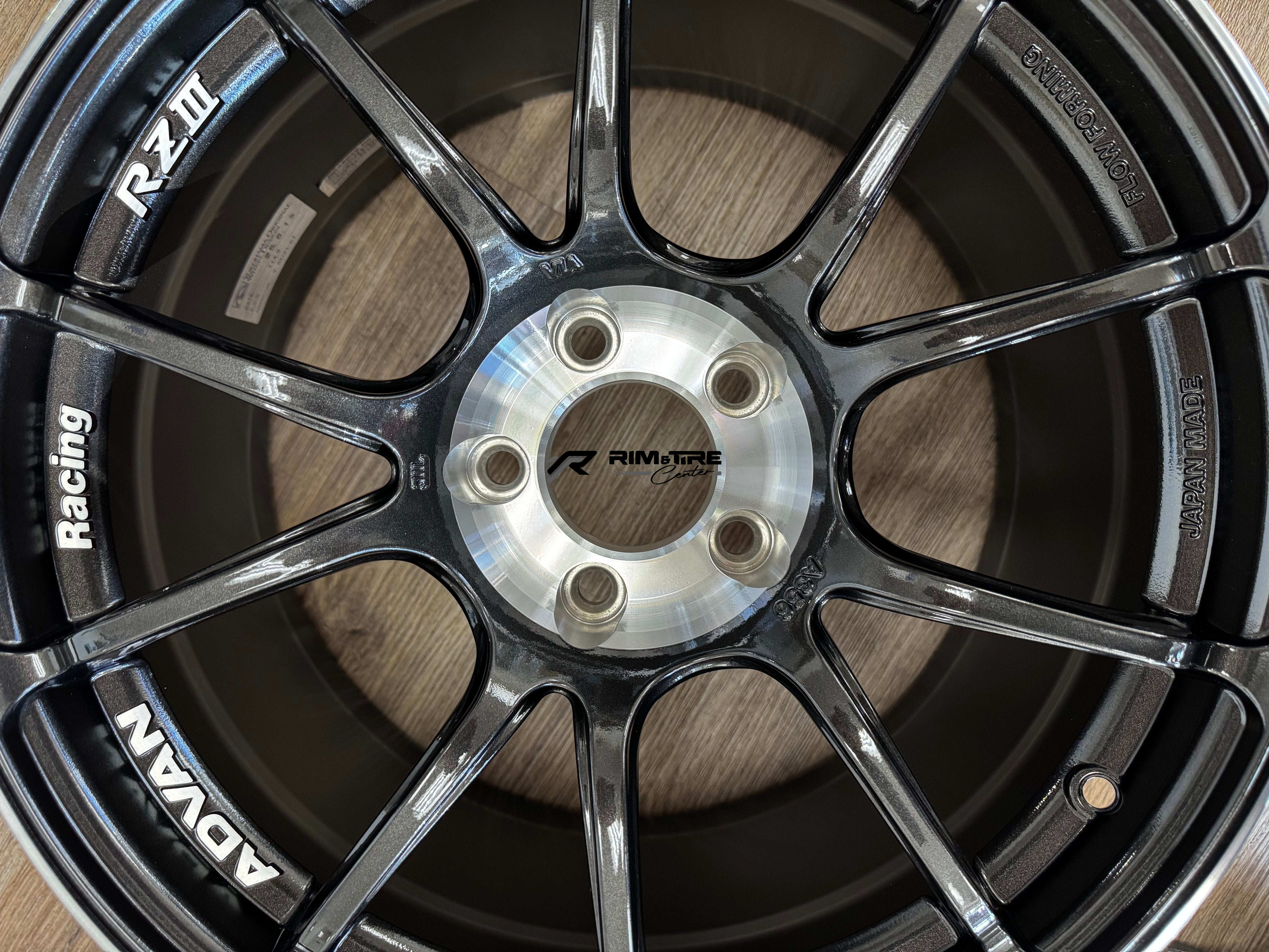 Advan Racing RZII dark metallic grey multi-spoke wheel with visible ADVAN and Racing branding, polished silver hub, and "Flow Forming" and "Japan Made" text on a wooden floor