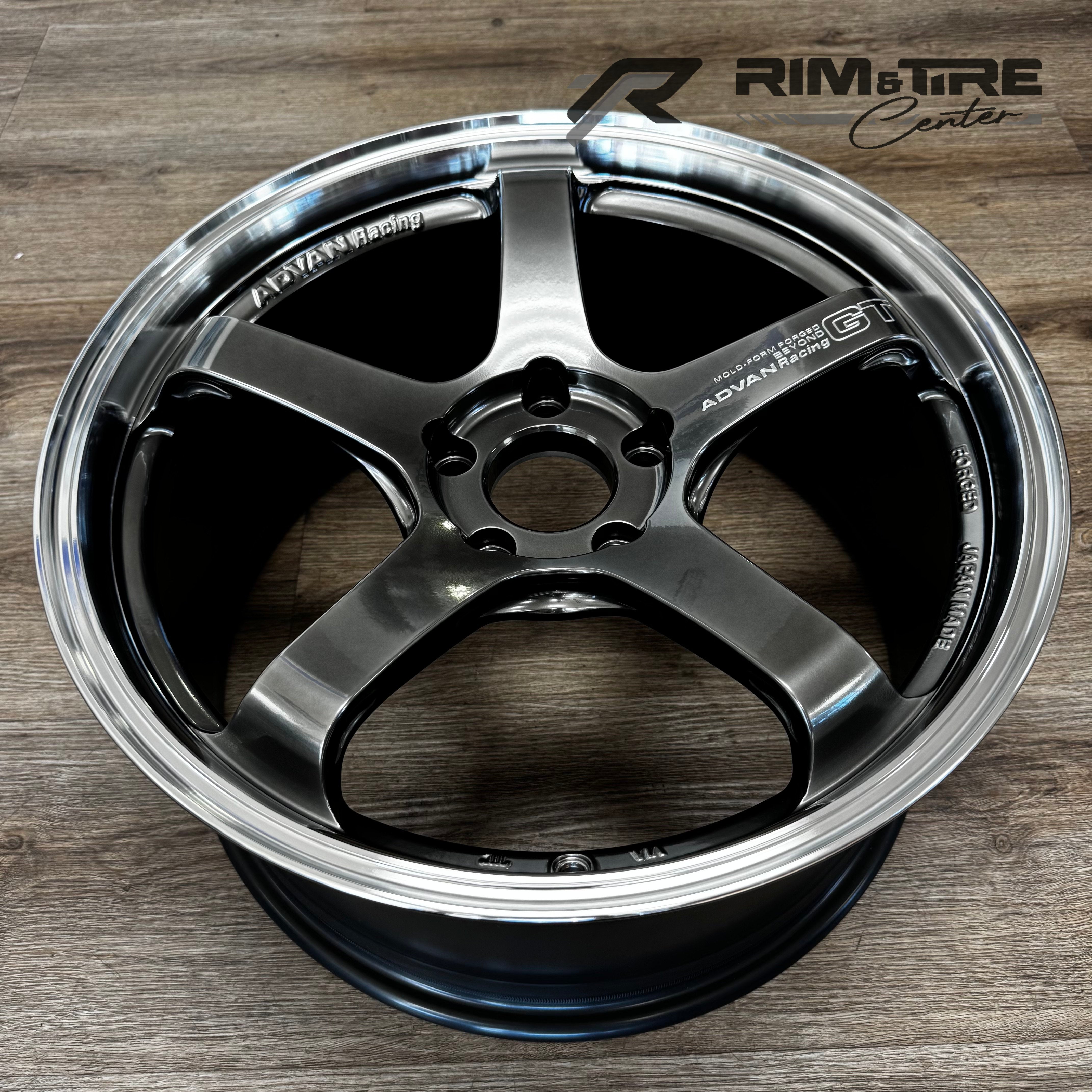 Photograph of an ADVAN Racing GT Beyond forged five-spoke dark metallic grey wheel rim with a polished chrome lip displayed on a light wooden floor showcasing "JAPAN MADE" text