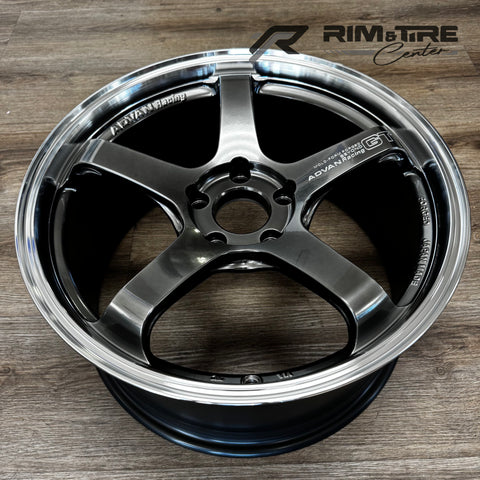 Photograph of an ADVAN Racing GT Beyond forged five-spoke dark metallic grey wheel rim with a polished chrome lip displayed on a light wooden floor showcasing "JAPAN MADE" text