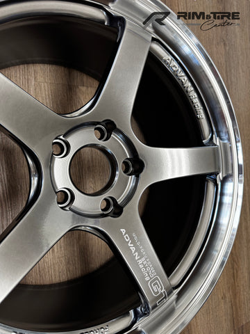 Photograph of a shiny gunmetal Advan Racing GT five-spoke forged car wheel with a polished chrome lip resting on a light brown wooden floor featuring prominent 'ADVAN Racing' text and a 'RIM & TIRE Center' watermark