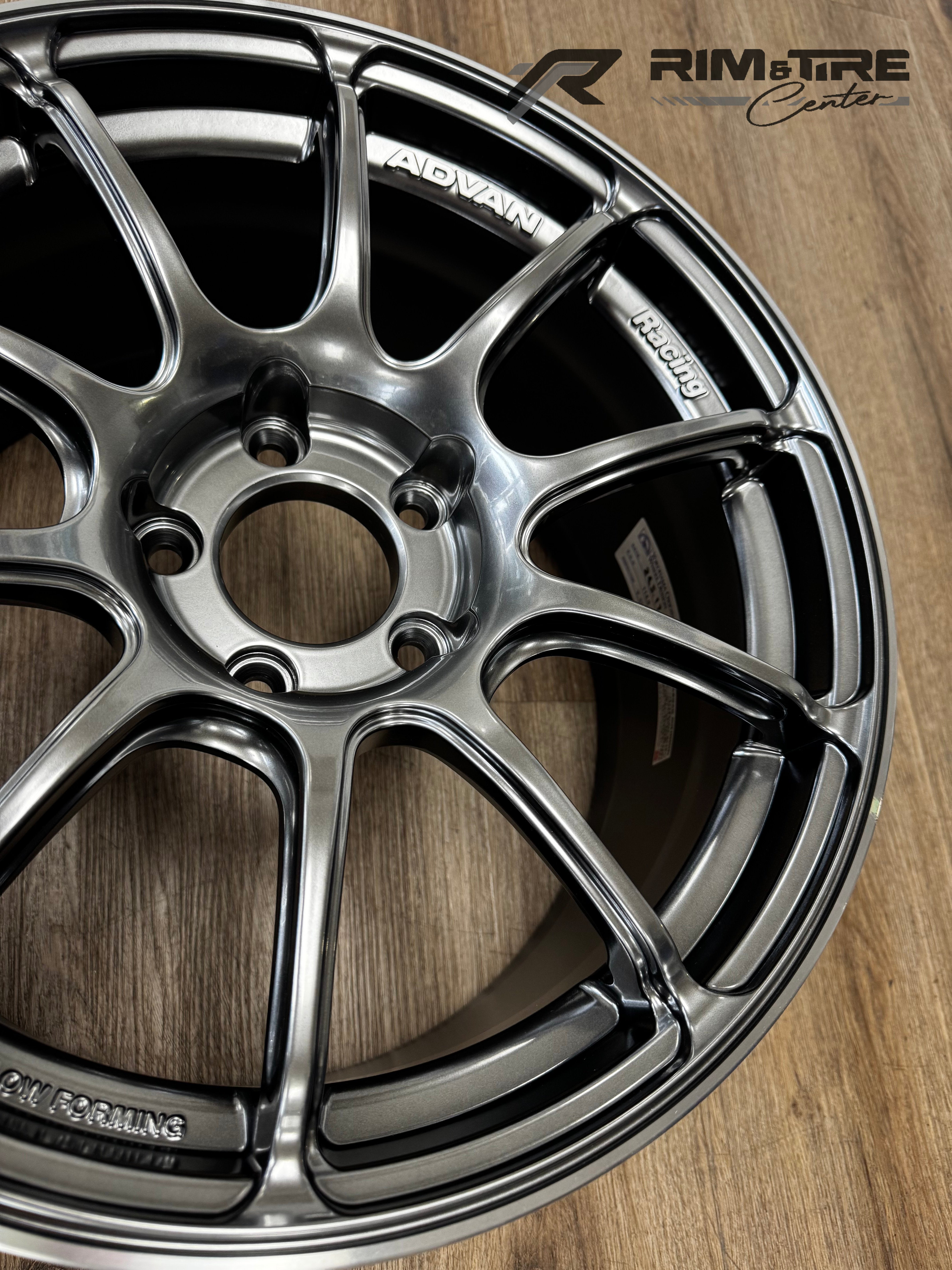Photography by RIM&TIRE Center of a metallic grey ADVAN Racing multi-spoke car wheel rim featuring 'ADVAN' and 'FLOW FORMING' text on a light wooden floor