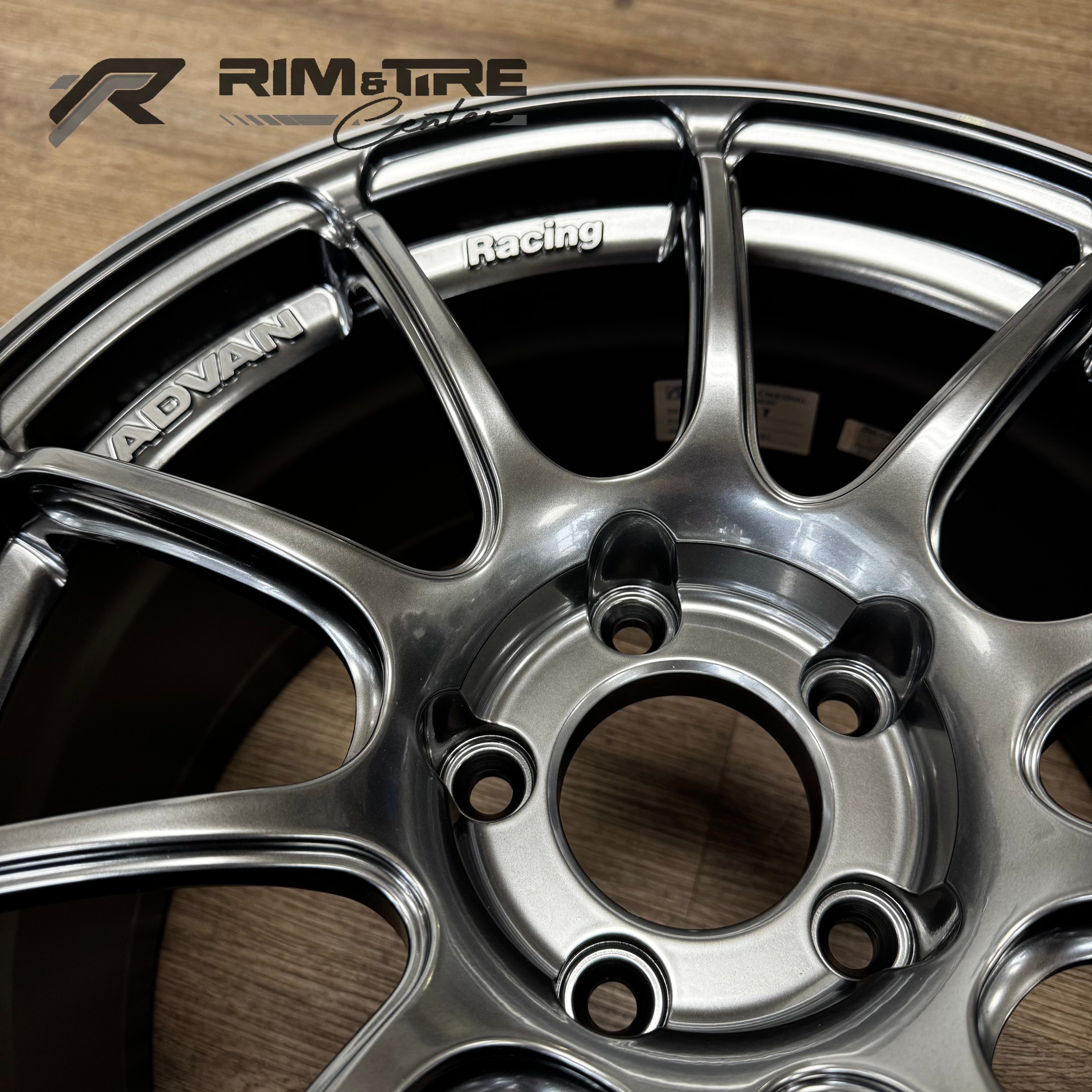 ADVAN Racing metallic silver multi-spoke car wheel photograph on a wooden floor showcasing ADVAN and Racing text, five black lug nut holes, and a RIM & TIRE Center logo