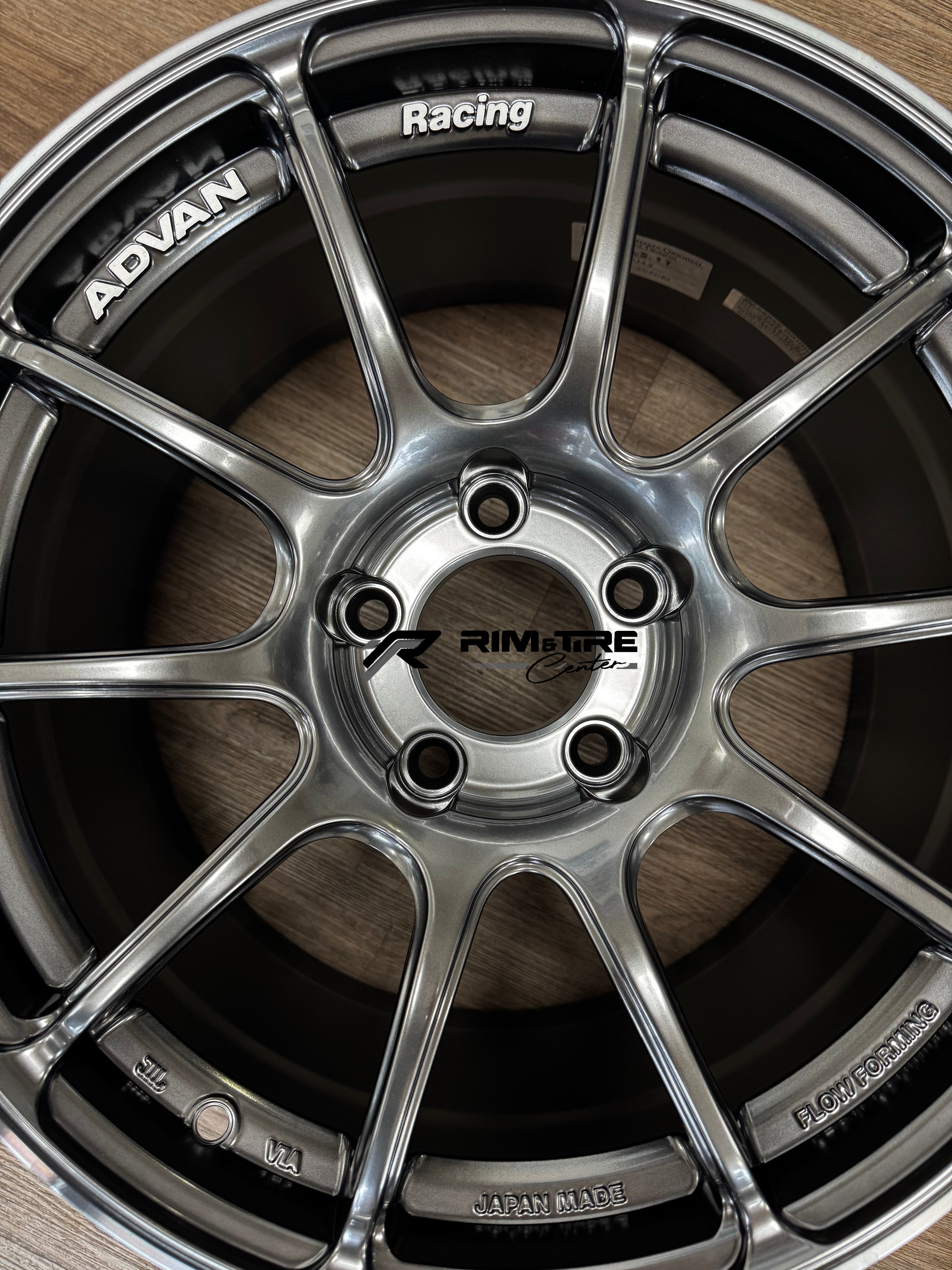 Photograph of an ADVAN Racing ten-spoke gunmetal car wheel on a wooden floor displaying JAPAN MADE and FLOW FORMING details and a Rim & Tire Center logo