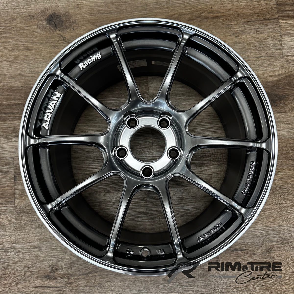 ADVAN Racing wheel, a dark gunmetal 10-spoke design with a polished lip, featuring "ADVAN", "Racing", and "JAPAN MADE" text, displayed on a light brown wooden floor